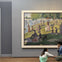 Two people sit and interact in front of a framed painting depicting a park scene with people by a riverside.