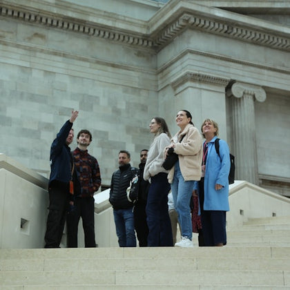 A group of people stand on steps inside a building with classical architecture, one person gesturing upwards.