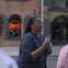 A person speaking to others outdoors near a brick building with arched windows and a neon sign that reads "Public Market."