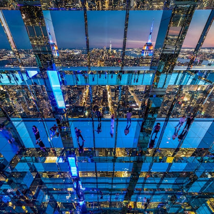 A mirrored interior reflecting people, city lights, and the skyline, including the Empire State Building at dusk.
