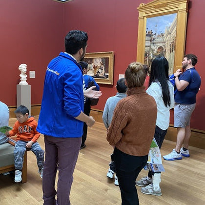 People observing art in a museum room with red walls. A guide wearing a blue jacket is speaking to a group. Busts and paintings are visible.