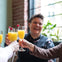 Four people clink glasses of orange drinks with raspberries. A person in the background is smiling, and there are plants near a window.