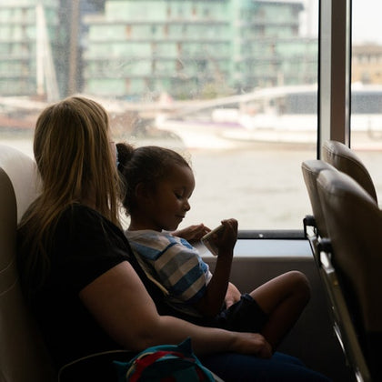 A woman and child sitting on a ferry, with the child looking at a phone and a cityscape visible through the window.