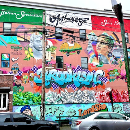 A vibrant mural covers a building's wall, featuring colorful graffiti, illustrations, text, and artistic portraits.