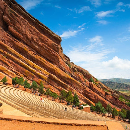 Outdoor amphitheater with large red rock formations, tiered seating, and scattered trees under a blue sky with clouds.