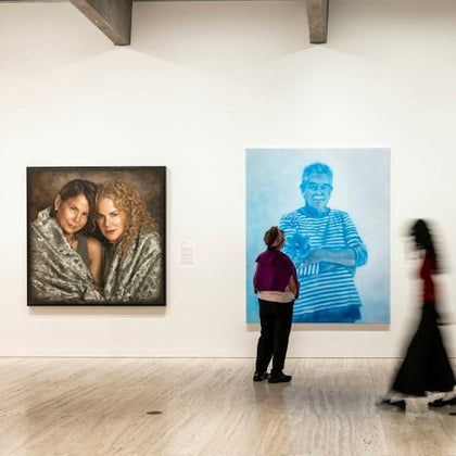 A gallery wall displaying two large paintings; one shows two women, the other a man. A person observes them closely.
