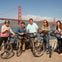 Five people with bicycles pose in front of the Golden Gate Bridge on a sunny day.