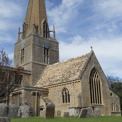A stone church with a tall spire and clock faces on its tower, surrounded by a graveyard under a partly cloudy sky.