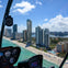 View from a helicopter cockpit overlooking a cityscape with tall buildings, a beach, and turquoise ocean under a blue sky.