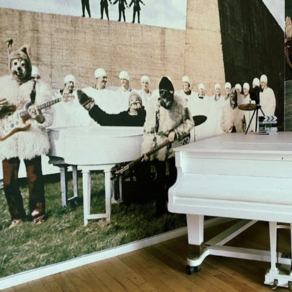A room with a white piano in front of a wall-sized mural depicting people in costumes and uniforms with musical instruments.