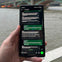 A hand holding a smartphone displaying a messaging app with a conversation visible. A river and bridge are in the background.