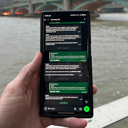 A hand holding a smartphone displaying a messaging app with a conversation visible. A river and bridge are in the background.