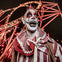 A person in clown makeup and costume standing in front of an illuminated roller coaster at night.
