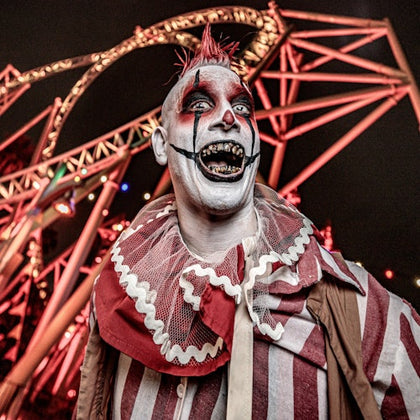 A person in clown makeup and costume standing in front of an illuminated roller coaster at night.