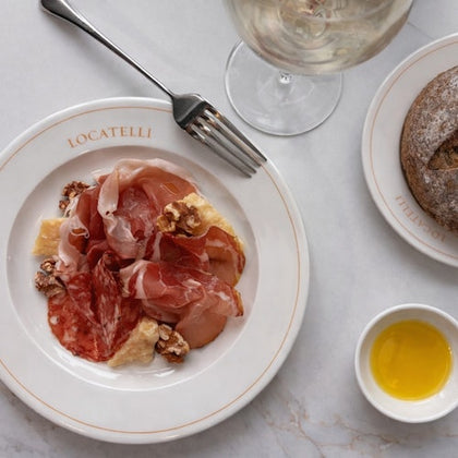 A plate with assorted cured meats, walnuts, and cheese, a loaf of bread, a glass of wine, and olive oil on a marble table.