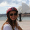 A smiling person wearing a red helmet and sunglasses outdoors with a blurred architectural structure in the background.