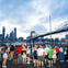 A crowd of people gather near the water, with the Brooklyn Bridge and New York City skyline in the background at dusk.