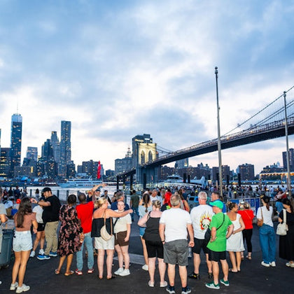 A crowd of people gather near the water, with the Brooklyn Bridge and New York City skyline in the background at dusk.