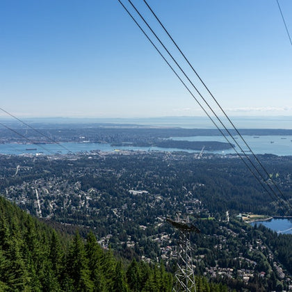 Aerial view of a cityscape with a forested hillside, power lines, and a body of water under a clear blue sky.