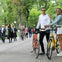 A man and a woman stand next to their bicycles in a park, with people walking and trees in the background.