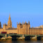 The Palace of Westminster with its gothic architecture, seen from across the Thames River under a clear blue sky.