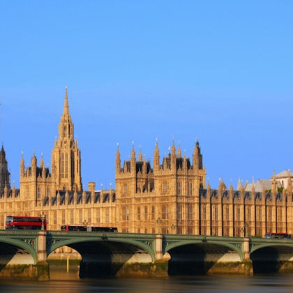 The Palace of Westminster with its gothic architecture, seen from across the Thames River under a clear blue sky.