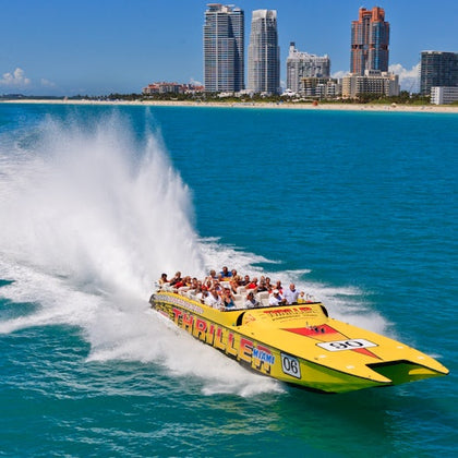 A speedboat full of people creates a large splash as it races through blue waters, with a cityscape visible in the background.