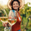 Two glasses of white wine held in focus, with a smiling woman wearing a hat blurred in the background in a vineyard.