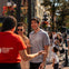 A guide in a red shirt interacts with smiling tourists in a lively urban area with flowers and pedestrians.