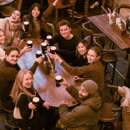 A group of people sitting around a wooden table in a cozy setting, holding up drinks and smiling cheerfully.