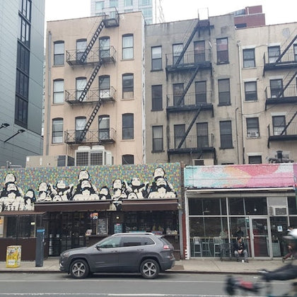 A city street with a mural of stormtroopers on a building, fire escapes on adjacent buildings, and a car driving by.