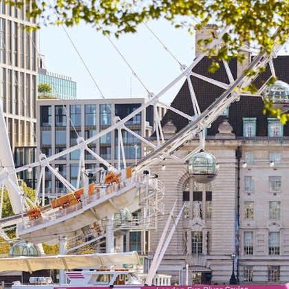 Urban scene with part of a Ferris wheel, glass capsules, modern buildings, and lush green trees. Boat on the river in the foreground.