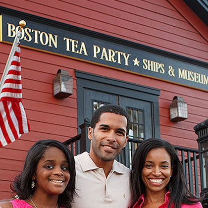 A group of three people smiling outside the Boston Tea Party Ships & Museum, with a U.S. flag and red building in the background.