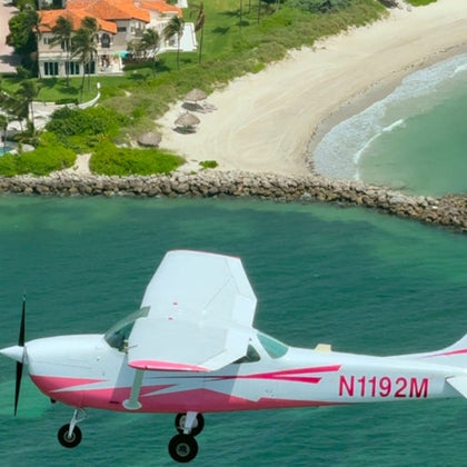 A white and pink propeller plane flies over turquoise water near a sandy beach with a house and palm trees in the background.