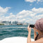 Person on a boat photographing a city skyline with a prominent tower, under a bright blue sky with scattered clouds.