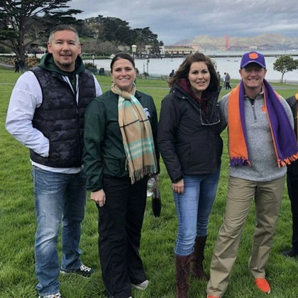 Five people standing on a grassy area with the Golden Gate Bridge and water in the background on a cloudy day.