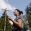 A person outdoors wearing casual clothing and glasses, gesturing with their hands, with a totem pole and trees in the background.