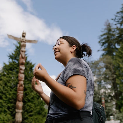A person outdoors wearing casual clothing and glasses, gesturing with their hands, with a totem pole and trees in the background.