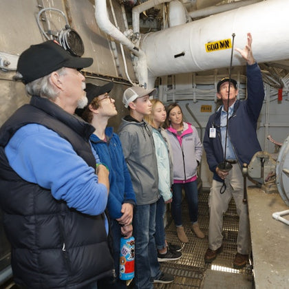A group of people stands inside a mechanical room while a guide gestures towards pipes and equipment.