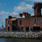 A brick building by a rocky waterfront, with the Statue of Liberty visible in the background under a partly cloudy sky.