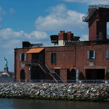 A brick building by a rocky waterfront, with the Statue of Liberty visible in the background under a partly cloudy sky.