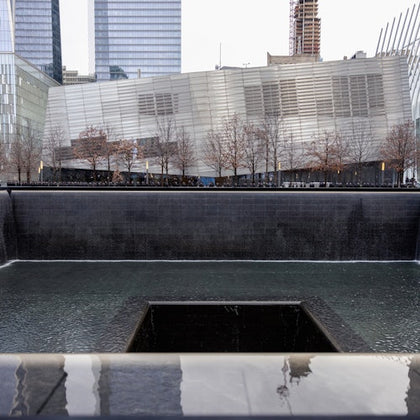 A reflecting pool with a central square void surrounded by a modern building and trees, within an urban setting.