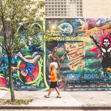 Person in an orange shirt walks past a colorful mural featuring various figures, bold text, and vibrant cosmic artwork.
