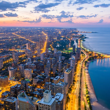Aerial view of a cityscape at dusk, with illuminated streets, tall buildings, and a shoreline along a calm body of water.