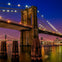 Brooklyn Bridge lit at night over calm water with a city skyline in the background and wooden posts in the foreground.