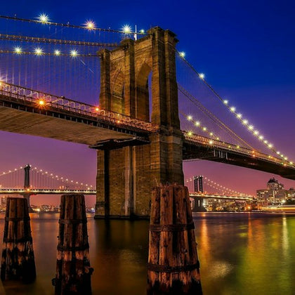 Brooklyn Bridge lit at night over calm water with a city skyline in the background and wooden posts in the foreground.