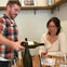 A man pours wine for a woman seated at a table with wine glasses and a tasting sheet in a cozy setting.