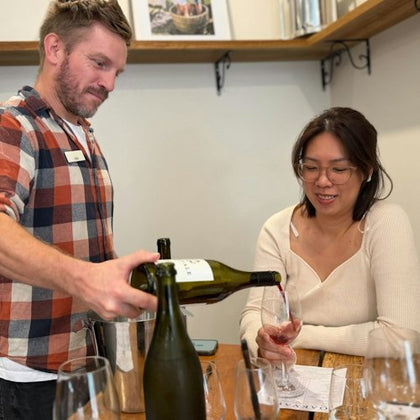 A man pours wine for a woman seated at a table with wine glasses and a tasting sheet in a cozy setting.