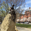 A bronze statue of a seated sailor atop a stone base in a park with blooming trees and buildings in the background.