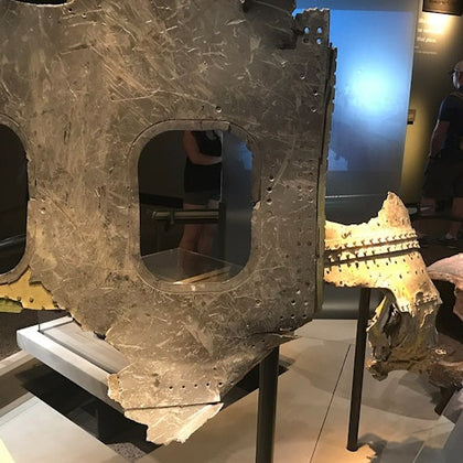A display of a damaged metal aircraft section with two window openings, supported by stands in a museum setting.